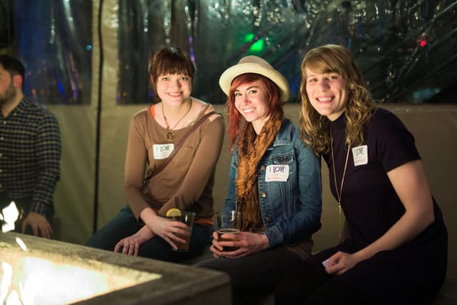 Three smiling women at a Love Bomb 2017 event, wearing name tags, seated near a fire pit, showcasing community engagement and support for My Voice Music nonprofit.